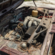 an under-hood photo of the engine bay of the burgundy 1974 bmw 2002tii. the car has sat abandoned in a field in florida for 25 years, and it shows: the engine compartment is full of leaves and branches and other trash, and the engine is covered in a thick layer of grime.